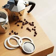 Close-up of three seals and one filter for 6-cup Bialetti Moka Express on wooden table; moka pot and coffee beans visible.