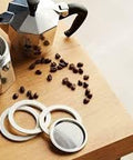 Close-up of stainless steel replacement filter and 3 gaskets for Moka Express 6-cup, on a wood table beside a moka pot.
