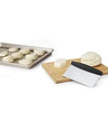 Stainless Steel All Purpose Scraper and Dough Cutter for Pizza Bread and Cookie Dough laying on cutting board beside dough on tray