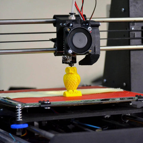 3D printer creating a small yellow owl figure on a print bed, showcasing precision in additive manufacturing.