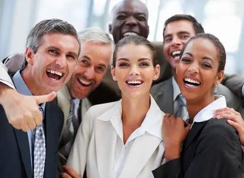 Group of smiling professionals in suits, celebrating teamwork and success in a bright office setting.