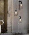 Industrial Style 60 Inch High Track Tree Floor Lamp With Iron Cage Three Lights Pipe LED Floor Light Fixture Black UL Listed E26 Base Bulbs Not Included Ideal For Living Rooms And Modern Spaces, positioned in a rustic room corner.