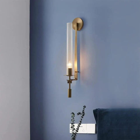 Modern Long Tube Wall Sconce In Brushed Gold Metal And Frosted Glass casting a warm glow on blue wall beside a bedside with decor.