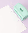 Mint finish 2 Hole Puncher With Waste Tray, 10 Sheet Capacity, by a sheet with punched holes, highlighting adjustable guide and non-slip base.