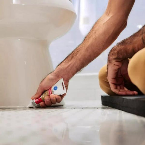 Person applying Advanced Silicone Caulk For Kitchen And Bathroom White 2.8 Fl Oz Tube 1 Pack to floor edge near a toilet on tiled surface.