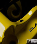 Prismatic Powders logo; close-up of glossy yellow automotive metal part with an arrow marker.