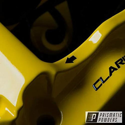 Prismatic Powders logo; close-up of glossy yellow automotive metal part with an arrow marker.
