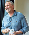 Smiling man holding a coffee mug while standing in a casual blue shirt by a window, enjoying a moment of relaxation.