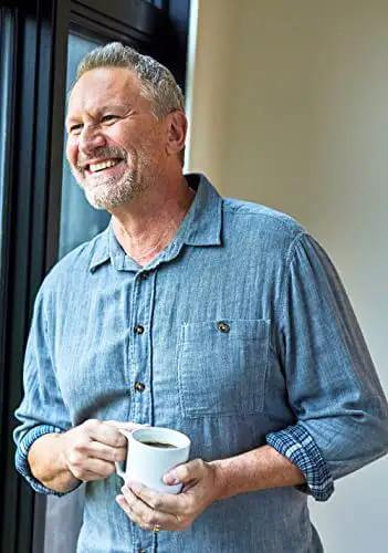 Smiling man holding a coffee mug while standing in a casual blue shirt by a window, enjoying a moment of relaxation.