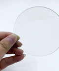 Hand holding a piece of the Pro Grade Lab Watch Glass Set 70mm Beaker Cover Lids For Chemistry 10 Pieces, clear and round glass disc.