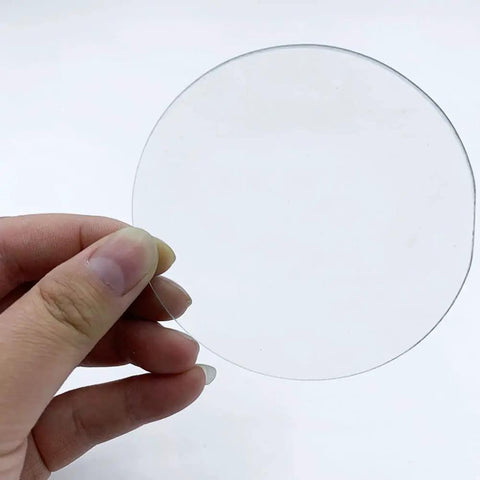 Hand holding a piece of the Pro Grade Lab Watch Glass Set 70mm Beaker Cover Lids For Chemistry 10 Pieces, clear and round glass disc.