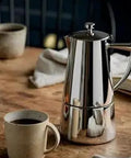 Stainless Steel Stovetop Espresso Maker For 6 Cups Easy To Clean Moka Pot With Heat Resistant Non Reactive Design Barista Grade Brew Durable Italian Style Coffee Percolator For Rich Bold Brew next to a coffee cup on a wooden table.