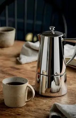 Stainless Steel Stovetop Espresso Maker For 6 Cups Easy To Clean Moka Pot With Heat Resistant Non Reactive Design Barista Grade Brew Durable Italian Style Coffee Percolator For Rich Bold Brew next to a coffee cup on a wooden table.