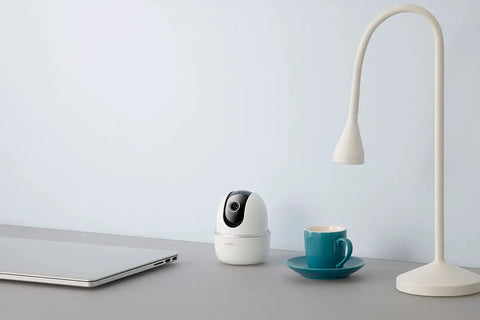 Indoor WiFi Security Camera With Pan And Tilt 4MP HD Sensor, placed on desk beside laptop, lamp, and cup
