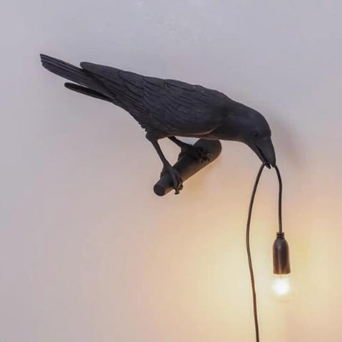 Crow Gothic Wall Lamp Sconce Bedside Bedroom And Living Room Raven Bird Night Light Left Facing Black Wall Lamp Farmhouse Decor Cozy Vibe Energy Saving Lit Mood Lighting with a hanging bulb held in a crow's beak.
