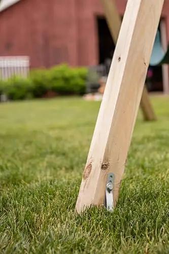 Close-up of a heavy-duty swing set ground anchor securing a wooden playset leg to the grass, part of Playset Anchor Kit For Swing Sets Heavy Duty Ground Anchors That Keep Your Set Rock Solid Stationary And Safer For Outdoor Fun Easy Install Swing Set Hardware To Secure Play Equipment And Protect Your Kids.