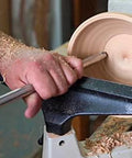 Front view of a woodturning lathe shaping a wooden bowl with a carbide-tipped tool and foam-grip handle