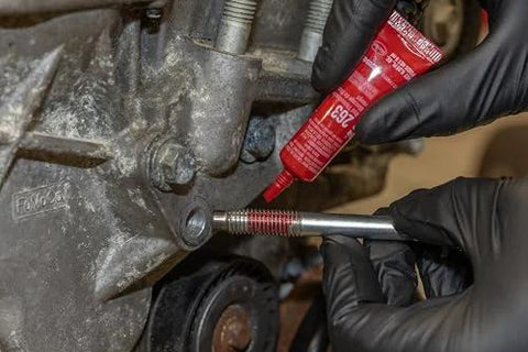 LOCTITE 263 red automotive threadlocker being applied to a bolt on an engine, close-up with gloved hands.