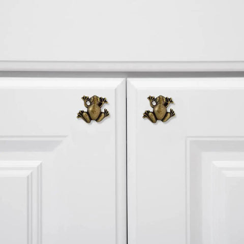 Two gold frog shaped cabinet handles on white cabinet doors, part of the Six Piece Frog Shaped Cabinet Handles Crafted From Solid Zinc Alloy Rust Proof Drawer Pulls And Dope Animal Knobs For Dresser Kitchen Cupboards Quick Mount Screws Included Fly Modern Home Decor Furniture Hardware.