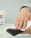 Person cleaning smartphone with SaniDry Disinfecting Wipes 300 Count EPA Approved Unscented White Nonabrasive Multi Surface Cleaning Wipes For Health Facilities Schools Industrial And Household Use Kills 99.9 Percent Of Viruses And Bacteria Including SARS CoV 2 And Influenza container on table.