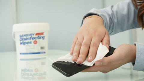 Person cleaning smartphone with SaniDry Disinfecting Wipes 300 Count EPA Approved Unscented White Nonabrasive Multi Surface Cleaning Wipes For Health Facilities Schools Industrial And Household Use Kills 99.9 Percent Of Viruses And Bacteria Including SARS CoV 2 And Influenza container on table.