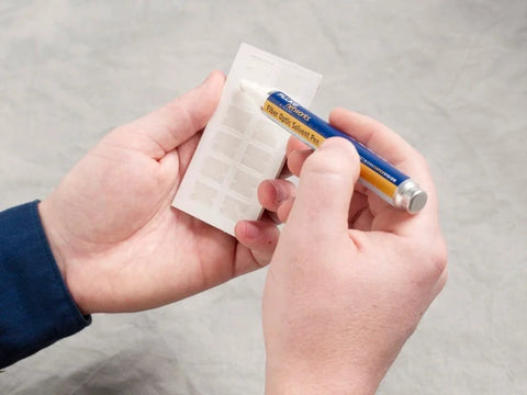 Fluke Networks NFC-SOLVENTPEN fiber optic cleaning solvent pen, blue body with yellow label, held in hands.