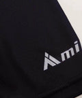 Close-up of black fabric with white "Ami" text logo, showcasing texture and seam detail.