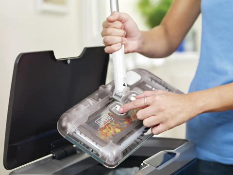 User operating No Fuss Grab And Go Cordless Floor Sweeper with swivel head over a dust bin for easy cleanup.