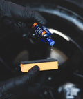 CARPRO CQUARTZ DLUX wheel and trim coat bottle (blue) held by gloved hand above orange sponge applicator.