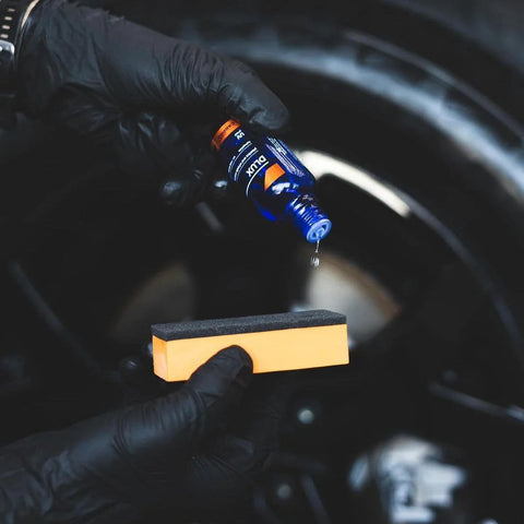 CARPRO CQUARTZ DLUX wheel and trim coat bottle (blue) held by gloved hand above orange sponge applicator.