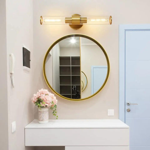 Gold Wall Sconce with Clear Glass in modern bathroom setting over round mirror, highlighting durable metal frame design.
