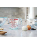 Prepware Glass Measuring Cup With Red Graphics, Clear, Non Porous Glass That Won't Stain Or Retain Odors, Microwave Safe, Oven Safe, And Dishwasher Safe, Great For Quick, Mess Free Kitchen Metrics. Clear glass measuring cup with red markings beside eggs and mixing bowl on kitchen counter