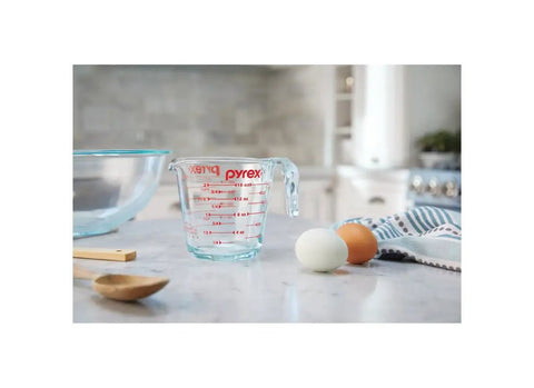 Prepware Glass Measuring Cup With Red Graphics, Clear, Non Porous Glass That Won't Stain Or Retain Odors, Microwave Safe, Oven Safe, And Dishwasher Safe, Great For Quick, Mess Free Kitchen Metrics. Clear glass measuring cup with red markings beside eggs and mixing bowl on kitchen counter