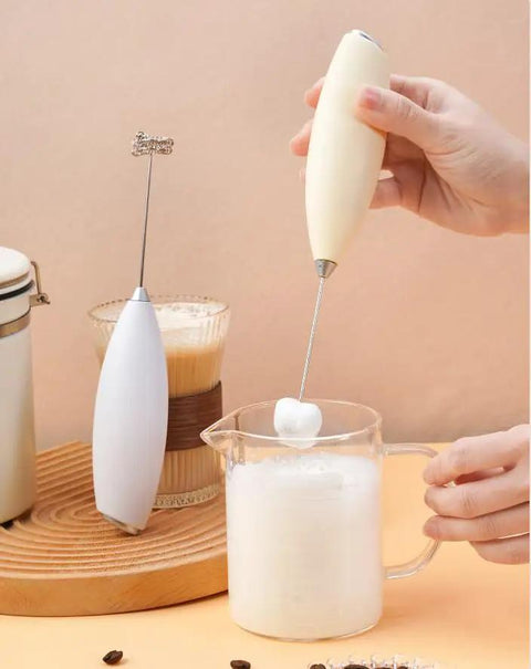 Handheld frother in use creating silky foam in a glass jug, part of Top Tier Handheld Milk Frother Packs Serious Oomph For Crafting Silky Foam On The Fly Ideal For Weekend Lattes Cappuccinos Matcha Mixes And Cozy Hot Chocolate Nights At Home.