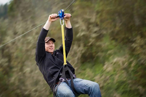 Man using 200 Ft Zip Line Kit With Adjustable Harness And Bungee Brake System for backyard fun, shown in action with blue trolley.