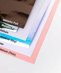Layers of metallic foil, vellum, adhesive vinyl, paper, and cardstock showcasing materials compatible with Alphanumeric Tiny Type Upper Thinlits Die Set 123 Pack For Cardmaking Journaling And Scrapbooking, Create Custom Titles With Intricate Cut Letters In Multicolor, Compatible With Paper Card Metallic Foil Vellum Shrink, By Tim Holtz Sizzix.