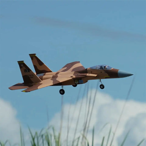 F-15 Twin 40mm EDF RC Fighter Jet PNP Desert Camo soaring above grass in flight with landing gear deployed, clear blue sky background.
