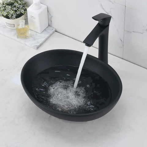 Oval dark vessel sink with black faucet and drain installed, water flowing into basin, modern design, ceramic, on marble counter