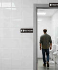 Two Pack Unisex Bathroom Signs For Doors Or Walls, Heavy Duty Metal Signs With Super Strong Adhesive, Easy Install For Home, Office Or Business Use, Black 9 Inch By 3 Inch displayed on bathroom door and wall with a person entering