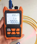 Orange handheld optical fiber tester with LCD readout and yellow fiber cable attached.