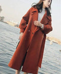 Woman wearing a long orange coat by the water, showcasing a stylish winter look with ankle boots.