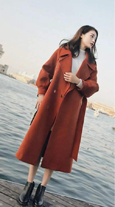 Woman wearing a long orange coat by the water, showcasing a stylish winter look with ankle boots.