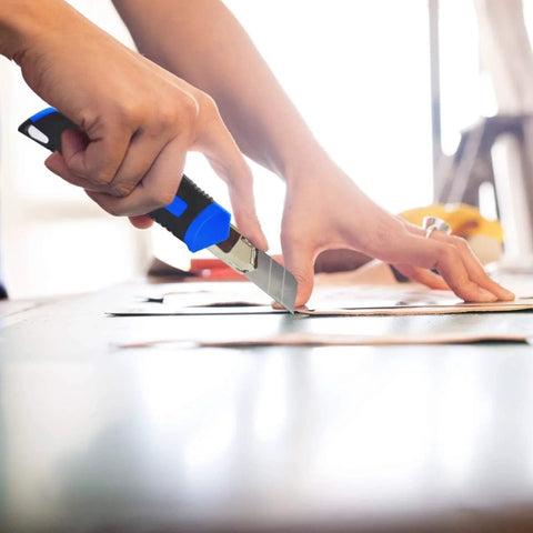 Close-up of a blue and black retractable utility knife cutting cardboard on a work surface