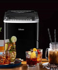Front view of Silonn countertop ice maker with black body and transparent lid, surrounded by drinks.