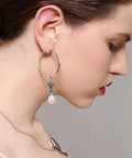 Woman wearing large hoop earrings with ornate design and dangling pearl.
