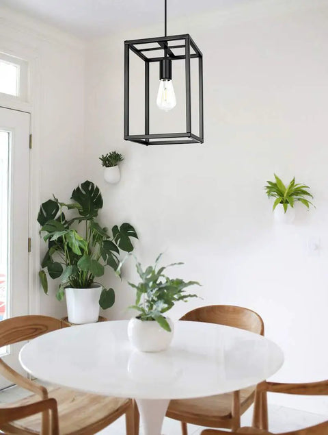 Industrial Black Pendant Light Fixture With Geometric Wire Cage hanging above a white dining table with plants in the background.