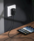 Universal 24W Multi Voltage AC DC Power Adapter plugged into an outlet, charging a tablet on a wooden table.