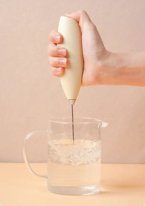 Handheld milk frother in action, blending water in a clear measuring cup, ideal for coffee drinks, Top Tier Handheld Milk Frother Packs Serious Oomph For Crafting Silky Foam On The Fly Ideal For Weekend Lattes Cappuccinos Matcha Mixes And Cozy Hot Chocolate Nights At Home.