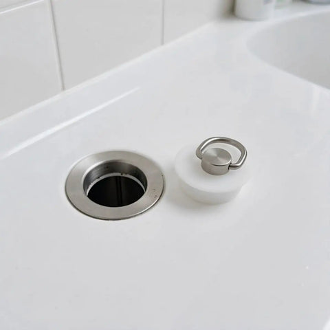 White Silicone Bathtub Water Stopper with Stainless Steel Ring placed next to a stainless steel sink drain.
