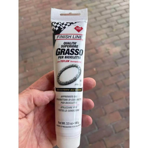 Heavy Duty Built Tough Bike Grease With High Pressure And Water Resistant Formula For Rust And Corrosion Protection Of Headsets Hubs And Bottom Brackets 3.5 Oz Ideal For Mountain And Road Bikes in a tube held by a hand on a red brick background